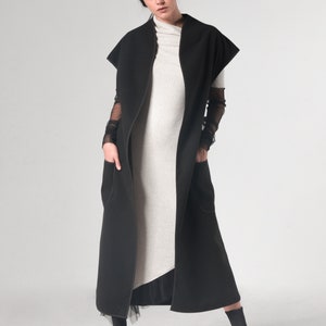 May include: A long black sleeveless coat with a high collar and pockets. The coat is worn over a light gray ribbed turtleneck dress.