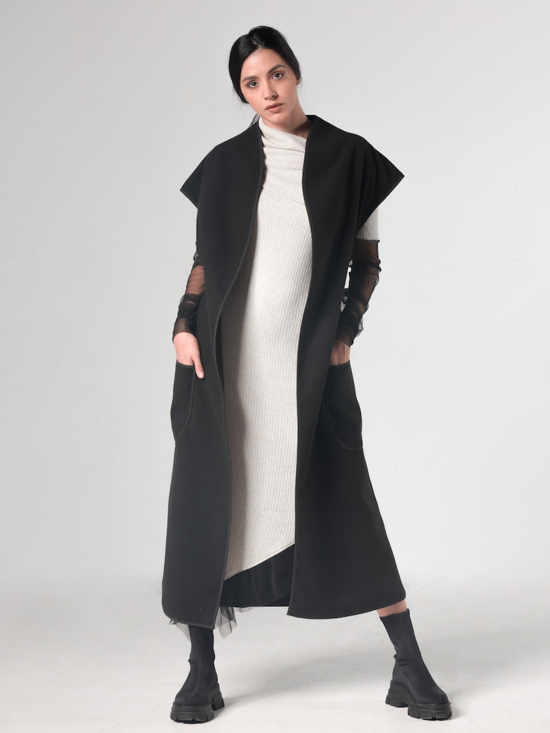 May include: A long black sleeveless coat with a high collar and pockets. The coat is worn over a light gray ribbed turtleneck dress.