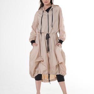 Puede incluir: Una chaqueta beige con capucha, cremallera negra y cintura con cordón. La chaqueta tiene mangas largas con puños negros y bolsillos grandes. La modelo lleva leggings negros y zapatos de plataforma.