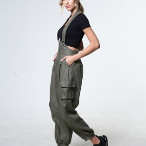 May include: A woman wearing a black crop top and olive green cargo overalls with adjustable straps. She is wearing black sneakers and has her hair pulled back in a ponytail.