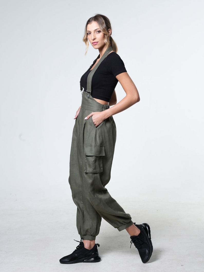 May include: A woman wearing a black crop top and olive green cargo overalls with adjustable straps. She is wearing black sneakers and has her hair pulled back in a ponytail.