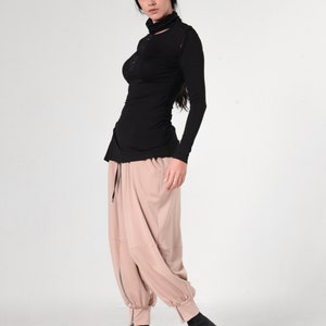 May include: A woman wearing a black long-sleeved shirt with a mock neck and a cut-out detail at the neckline. She is also wearing beige pants with a drawstring waist and a zipper at the ankles. She is wearing black ankle boots.