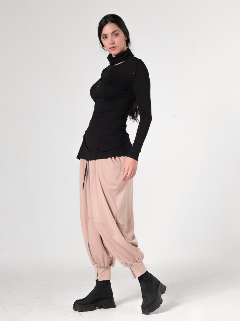 May include: A woman wearing a black long-sleeved shirt with a mock neck and a cut-out detail at the neckline. She is also wearing beige pants with a drawstring waist and a zipper at the ankles. She is wearing black ankle boots.