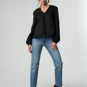 Puede incluir: Una mujer con una blusa negra de manga larga con cuello en V y jeans azules rectos. Lleva zapatos negros de tac&oacute;n.