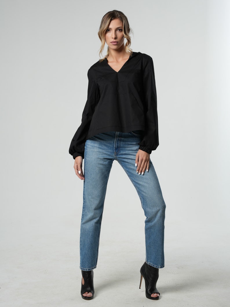 Puede incluir: Una mujer con una blusa negra de manga larga con cuello en V y jeans azules rectos. Lleva zapatos negros de tac&oacute;n.
