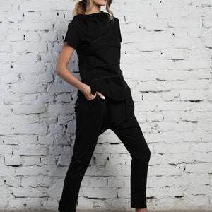 May include: A woman wearing a black, layered, short-sleeved top and black pants with a loose fit around the ankles. She is wearing white sandals with straps.
