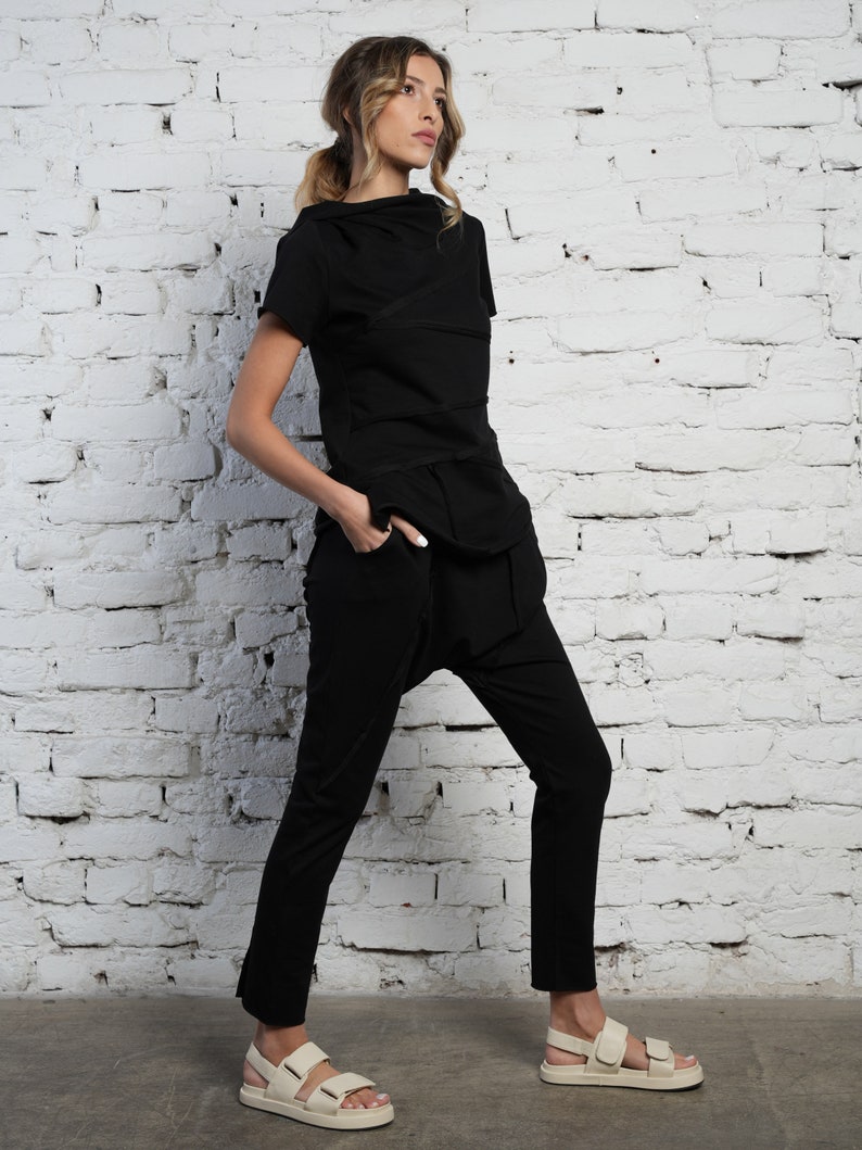 May include: A woman wearing a black, layered, short-sleeved top and black pants with a loose fit around the ankles. She is wearing white sandals with straps.