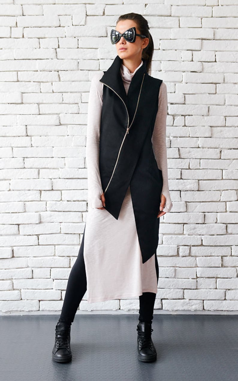 May include: A black sleeveless jacket with a zipper closure and a high collar. The jacket is worn over a long-sleeved white shirt and black leggings. The person is wearing black high-top sneakers and oversized black sunglasses.