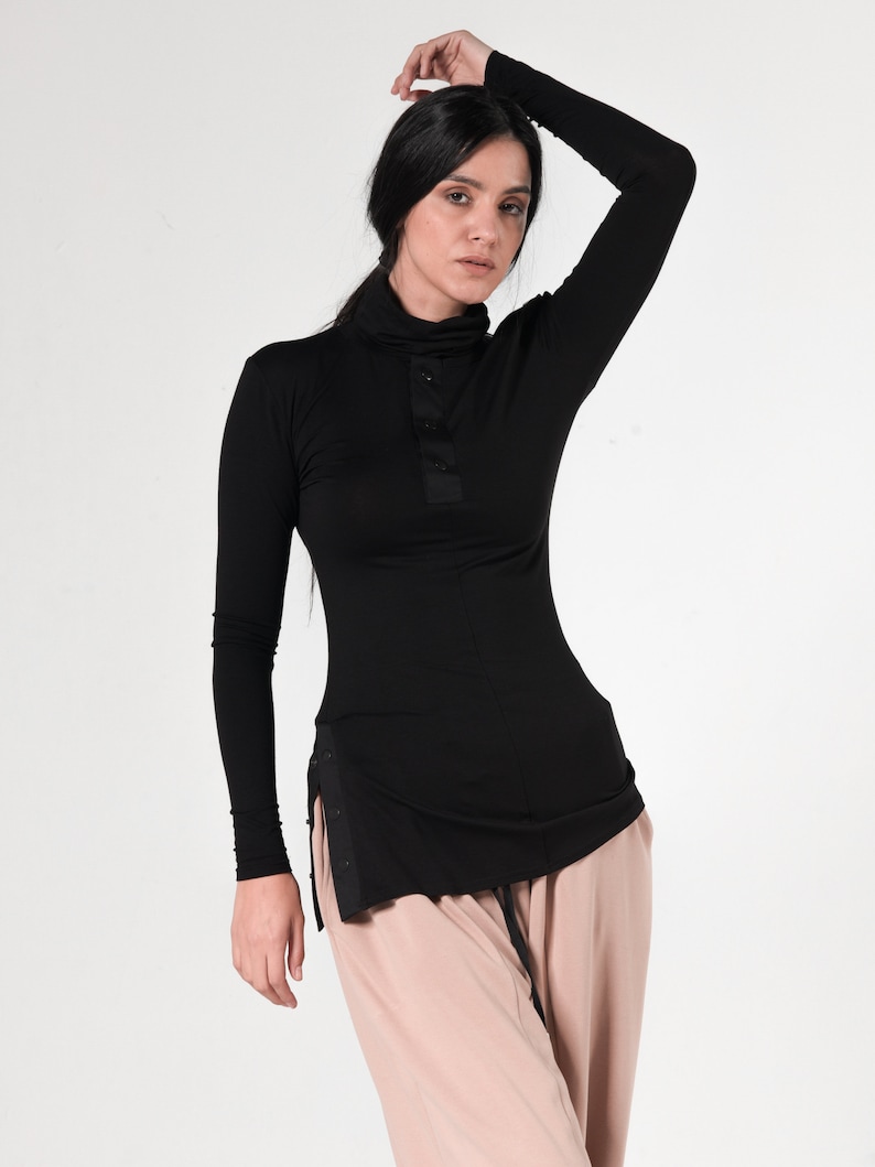 May include: A black long-sleeved shirt with a mock neck and a single button closure. The shirt has a side slit and a slightly longer length in the back.