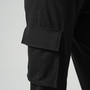 May include: Black cargo pants with a large, rectangular pocket on the left leg. The pocket has a flap and is sewn with a contrasting stitch.