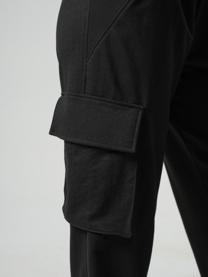 May include: Black cargo pants with a large, rectangular pocket on the left leg. The pocket has a flap and is sewn with a contrasting stitch.
