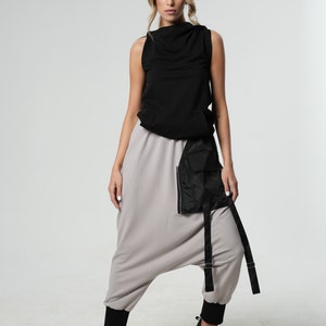 May include: A woman is wearing a black sleeveless top with a cowl neck and gray harem pants with a black utility belt. She is wearing black sneakers.