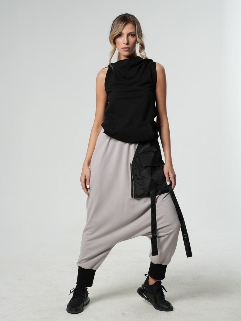 May include: A woman is wearing a black sleeveless top with a cowl neck and gray harem pants with a black utility belt. She is wearing black sneakers.