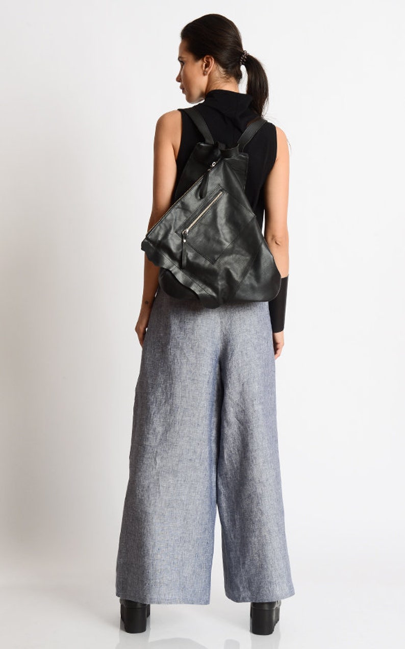 May include: A black leather backpack with a zippered pocket and a flap closure. The backpack is being worn by a person wearing a black sleeveless shirt and blue linen pants.