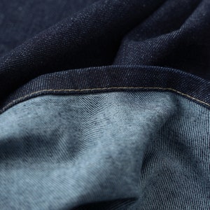 Puede incluir: Primer plano de tela vaquera azul oscuro con una costura visible. La tela est&aacute; doblada y texturizada, mostrando el tejido del material.