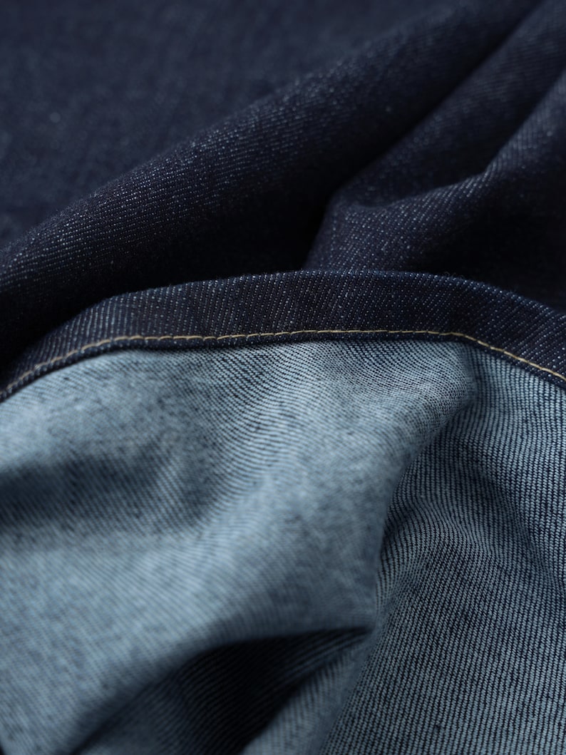 Puede incluir: Primer plano de tela vaquera azul oscuro con una costura visible. La tela est&aacute; doblada y texturizada, mostrando el tejido del material.