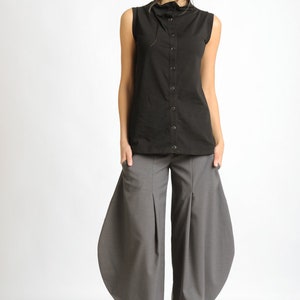 May include: A woman is wearing a black sleeveless shirt with a high collar and a button-up front. She is also wearing gray pants with a wide leg and a gathered design. The pants have pockets on the sides. She is wearing black shoes with white soles.