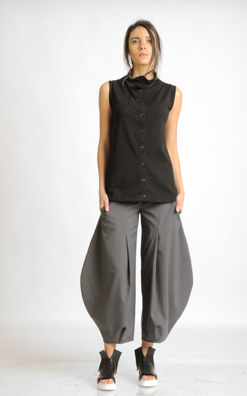 May include: A woman is wearing a black sleeveless shirt with a high collar and a button-up front. She is also wearing gray pants with a wide leg and a gathered design. The pants have pockets on the sides. She is wearing black shoes with white soles.