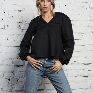 May include: A black long-sleeved top with a V-neck and gathered sleeves, worn with light blue denim jeans. The model is standing in front of a white brick wall.