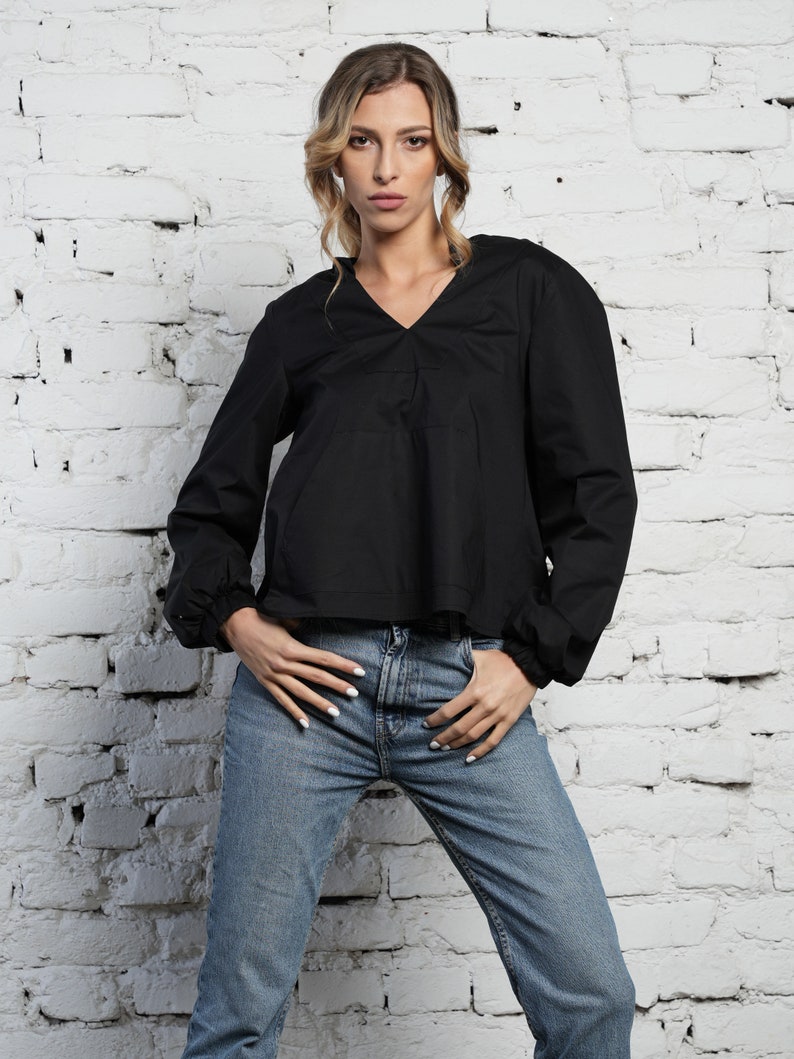 May include: A black long-sleeved top with a V-neck and gathered sleeves, worn with light blue denim jeans. The model is standing in front of a white brick wall.
