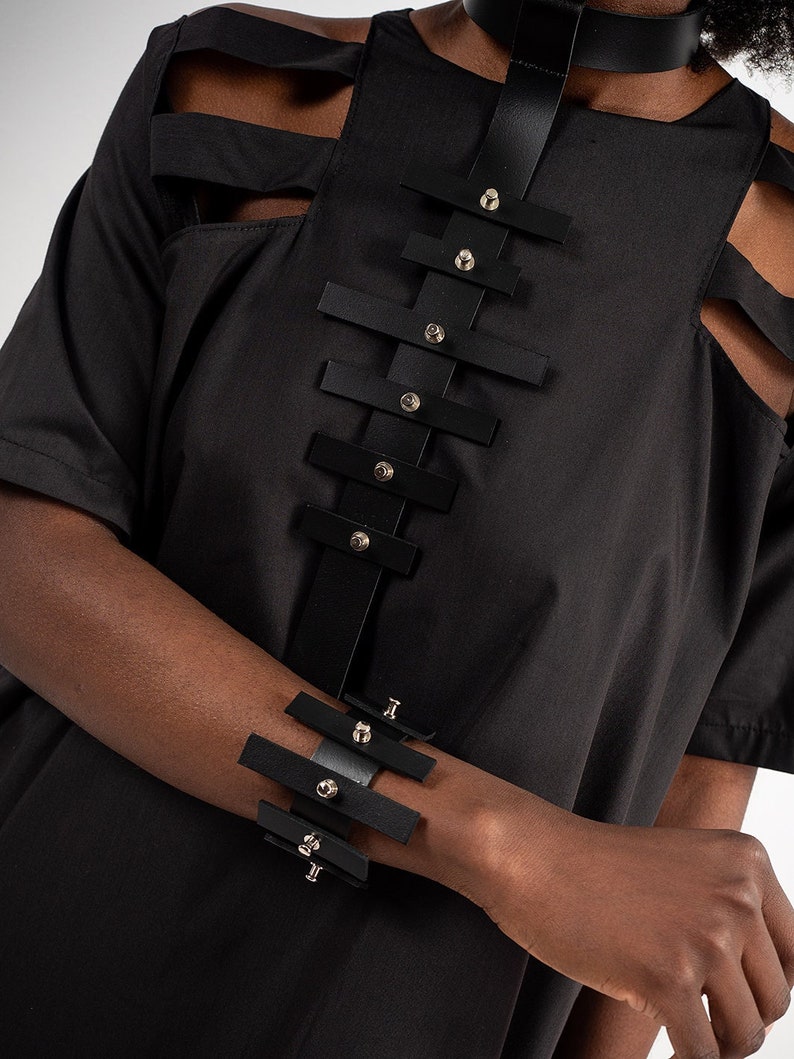 Puede incluir: Un collar gargantilla y una pulsera negros con tachuelas plateadas y m&uacute;ltiples correas. Las correas est&aacute;n conectadas por anillos met&aacute;licos.