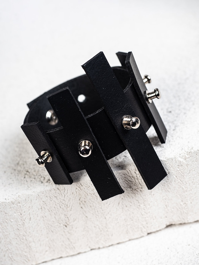 Puede incluir: Pulsera de cuero negro con un dise&ntilde;o geom&eacute;trico y detalles en metal plateado.