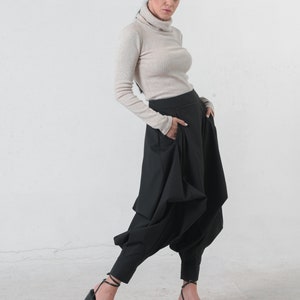 May include: A woman wearing a beige turtleneck sweater and black pants with a unique, gathered design. She is wearing black heeled sandals.