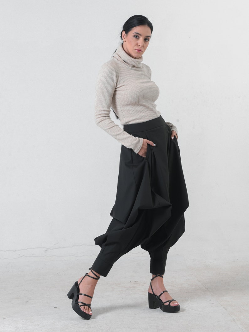 May include: A woman wearing a beige turtleneck sweater and black pants with a unique, gathered design. She is wearing black heeled sandals.