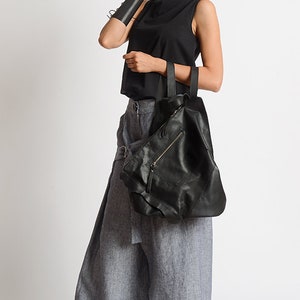 May include: A woman wearing a black sleeveless top, gray linen pants, and black platform shoes. She is carrying a large black leather tote bag with a zippered pocket.