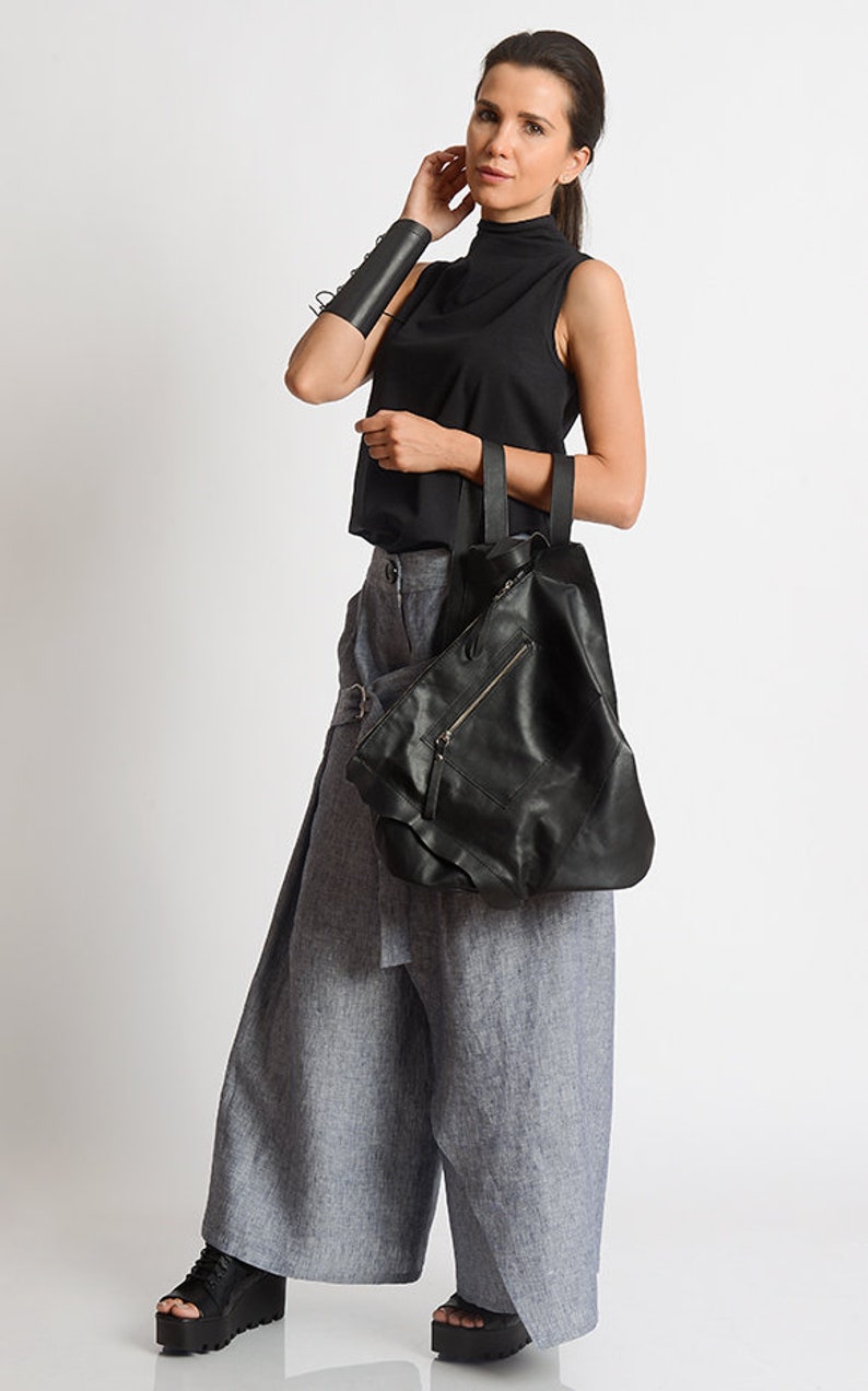 May include: A woman wearing a black sleeveless top, gray linen pants, and black platform shoes. She is carrying a large black leather tote bag with a zippered pocket.