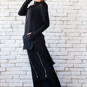 May include: A black hooded sweatshirt with a long, asymmetrical skirt. The skirt has multiple zippers down the front. The model is wearing white sneakers.