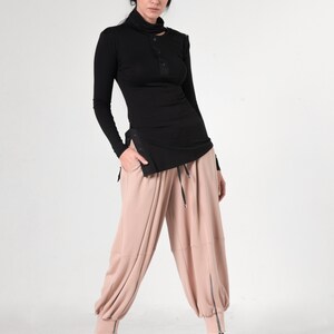 May include: A woman is wearing a black mock neck shirt with a single button detail and a pair of loose-fitting, blush-colored pants with zipper accents at the ankles.