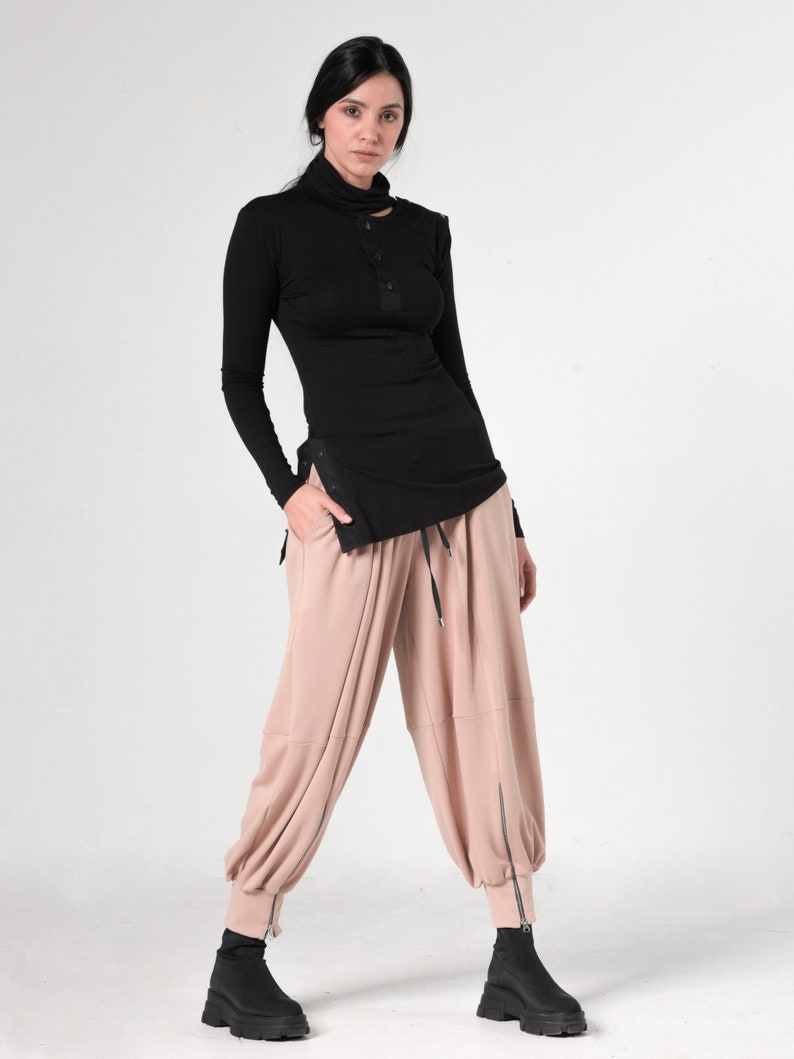May include: A woman is wearing a black mock neck shirt with a single button detail and a pair of loose-fitting, blush-colored pants with zipper accents at the ankles.