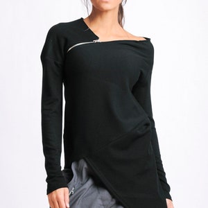 Könnte beinhalten: Schwarzes Langarmshirt mit einem diagonalen Reißverschluss und einem asymmetrischen Saum. Das Shirt wird mit einer grauen Hose kombiniert.