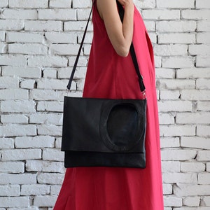 May include: A woman wearing a bright red sleeveless maxi dress and black sunglasses is carrying a black leather satchel with a circular cutout. The bag has a long black strap.