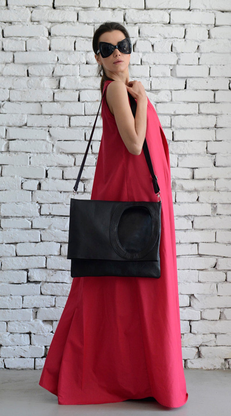 May include: A woman wearing a bright red sleeveless maxi dress and black sunglasses is carrying a black leather satchel with a circular cutout. The bag has a long black strap.