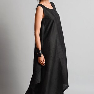 May include: A black sleeveless linen dress with a flowing, asymmetrical hemline. The dress has a simple, round neckline and a single pocket on the left side. The model is wearing black sneakers.