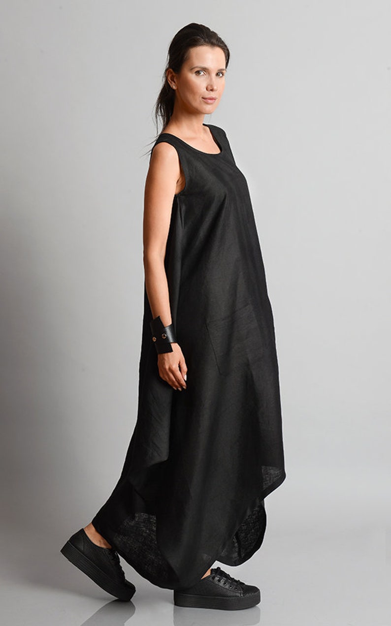 May include: A black sleeveless linen dress with a flowing, asymmetrical hemline. The dress has a simple, round neckline and a single pocket on the left side. The model is wearing black sneakers.