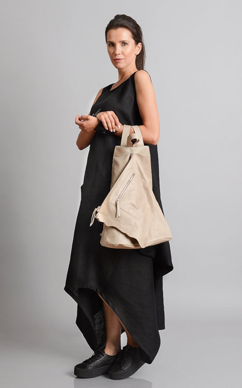 May include: A woman wearing a black sleeveless dress with a high low hemline and a beige leather bag. She is wearing black sneakers.