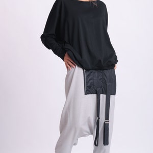 May include: A woman wearing a black long-sleeved top and light gray harem pants. The pants have a black panel with a zipper and black straps. Black shoes complete the outfit. The clothing is modern and stylish.
