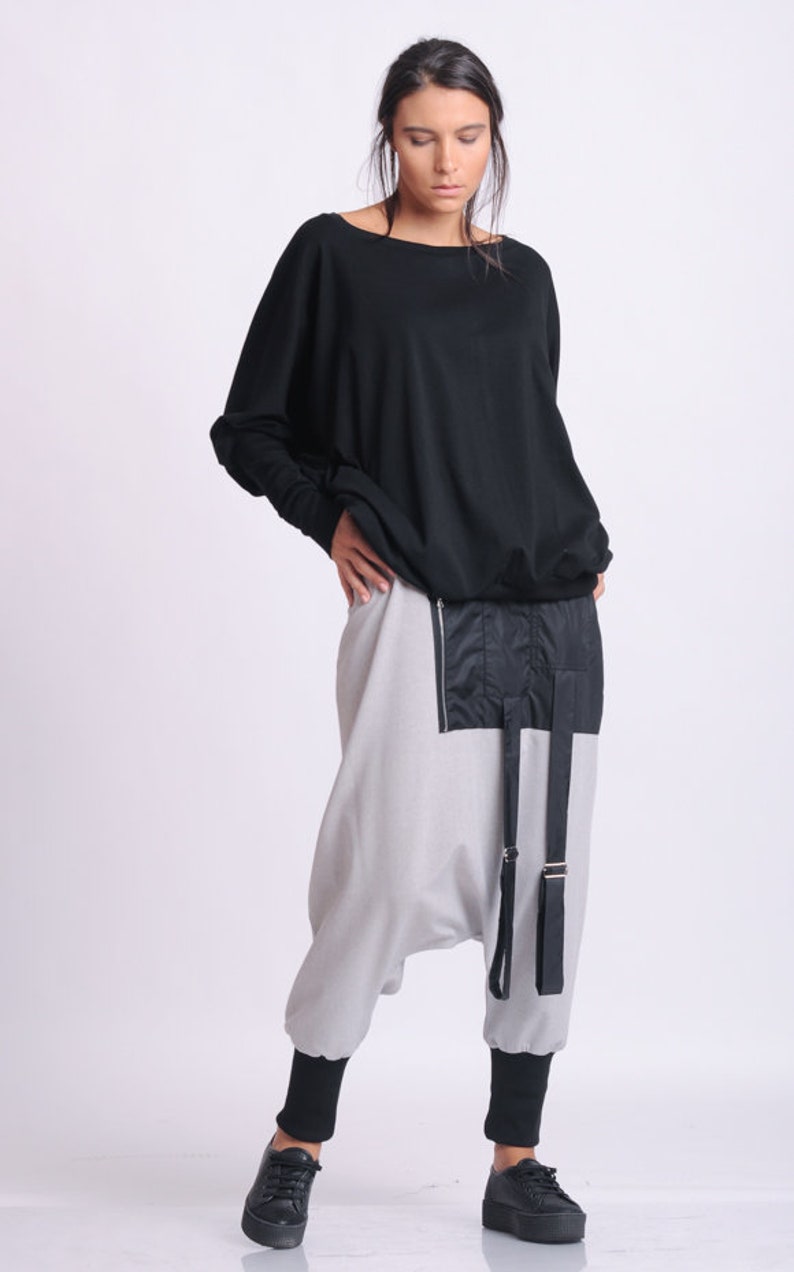 May include: A woman wearing a black long-sleeved top and light gray harem pants. The pants have a black panel with a zipper and black straps. Black shoes complete the outfit. The clothing is modern and stylish.