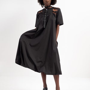 May include: A black midi dress with a unique design featuring a leather strap detail across the chest. The dress has a loose, flowing silhouette and short sleeves.