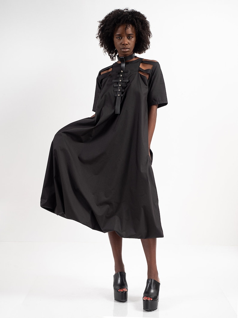 May include: A black midi dress with a unique design featuring a leather strap detail across the chest. The dress has a loose, flowing silhouette and short sleeves.
