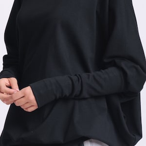 May include: Black long-sleeved top with a wide neckline and a relaxed fit. The sleeves are fitted at the wrists. The bottom of the top has a gathered, slightly flared design. The model is wearing black and gray pants.
