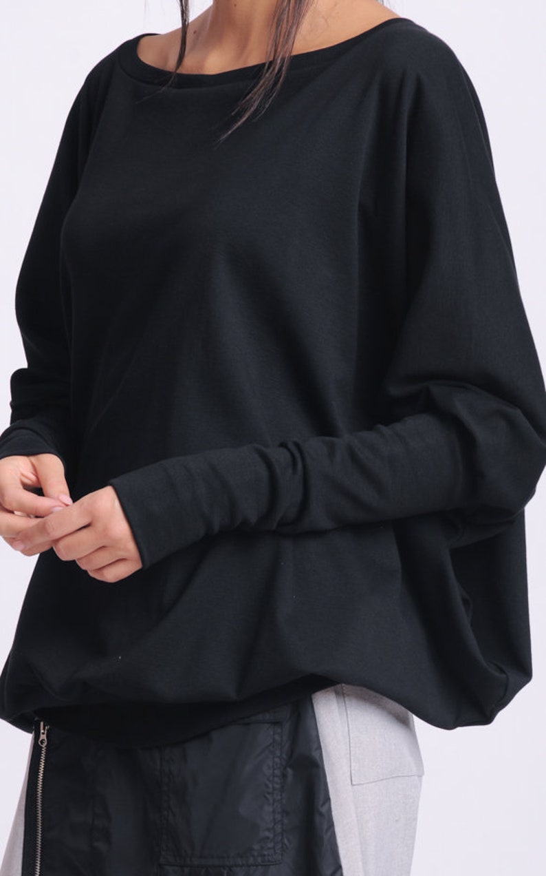 May include: Black long-sleeved top with a wide neckline and a relaxed fit. The sleeves are fitted at the wrists. The bottom of the top has a gathered, slightly flared design. The model is wearing black and gray pants.