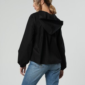 May include: A black hooded jacket with long sleeves and a relaxed fit. The jacket is paired with light blue denim jeans. The model is seen from the back, against a neutral background.
