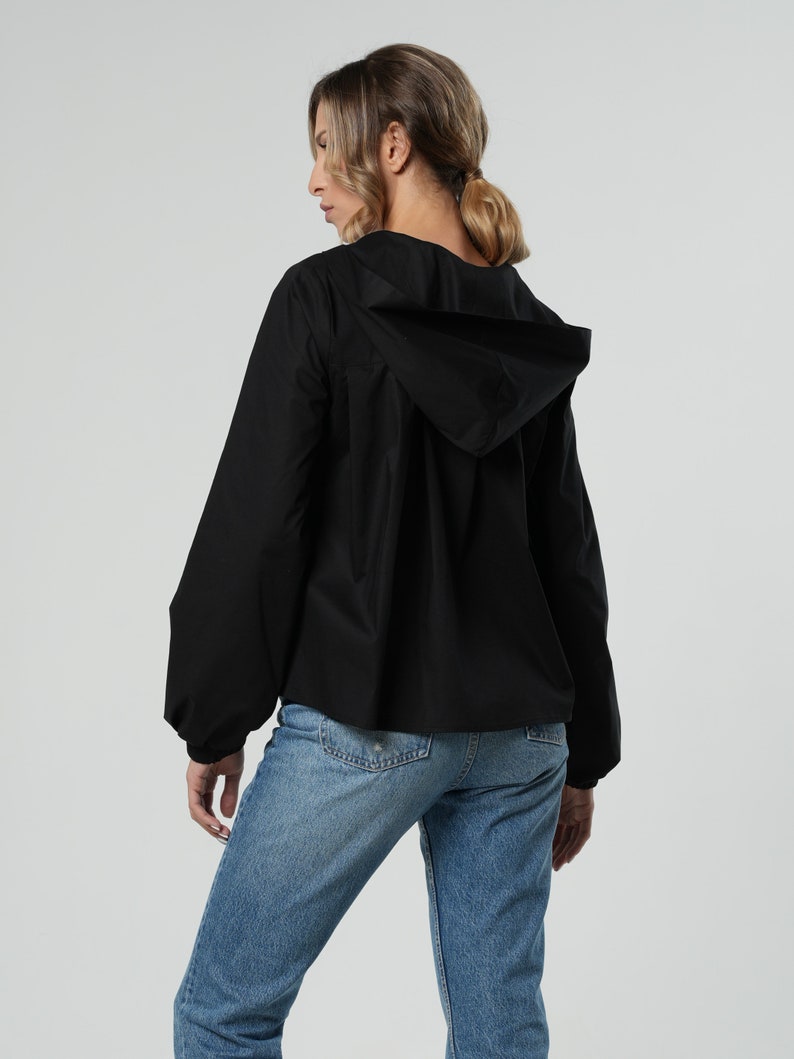 May include: A black hooded jacket with long sleeves and a relaxed fit. The jacket is paired with light blue denim jeans. The model is seen from the back, against a neutral background.