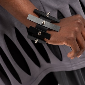 Puede incluir: Un brazalete de cuero negro y gris con tachuelas plateadas. El brazalete est&aacute; hecho de m&uacute;ltiples tiras de cuero que se superponen y se conectan con remaches met&aacute;licos.