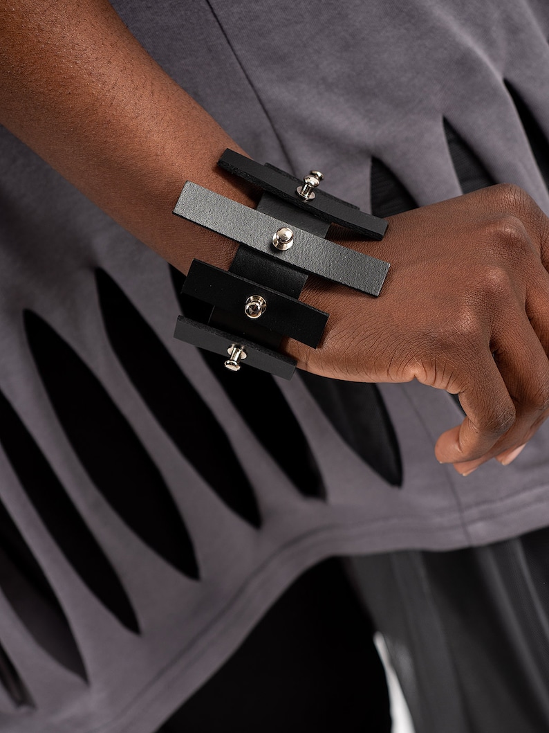 Puede incluir: Un brazalete de cuero negro y gris con tachuelas plateadas. El brazalete est&aacute; hecho de m&uacute;ltiples tiras de cuero que se superponen y se conectan con remaches met&aacute;licos.