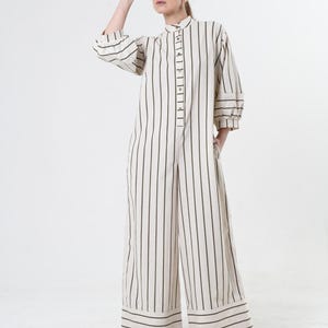 May include: A white and black striped jumpsuit with a button-up front and long sleeves. The jumpsuit has a wide leg and a relaxed fit. The model is wearing a light brown cap.